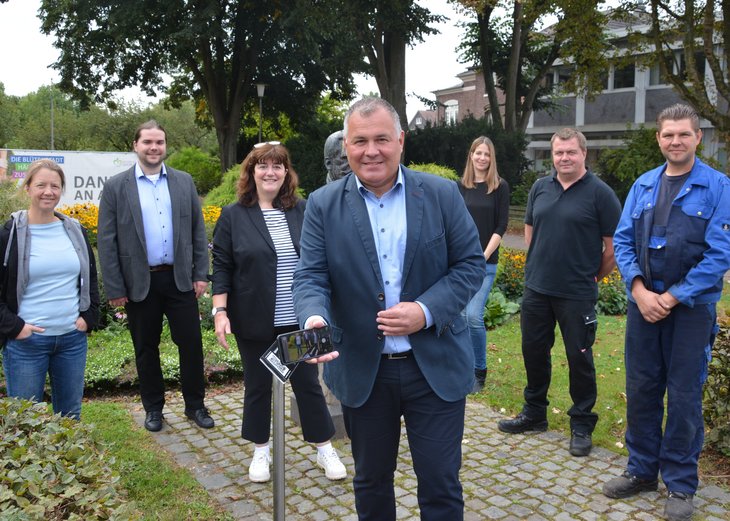 Bürgermeister Steffes steht vor der Julius-Pohlig-Büste, sein Handy liest den QR-Code der Stele. Im Hintergrund steht das Team der Stadtverwaltung, das die digitale Stadtführung entwickelt hat. 