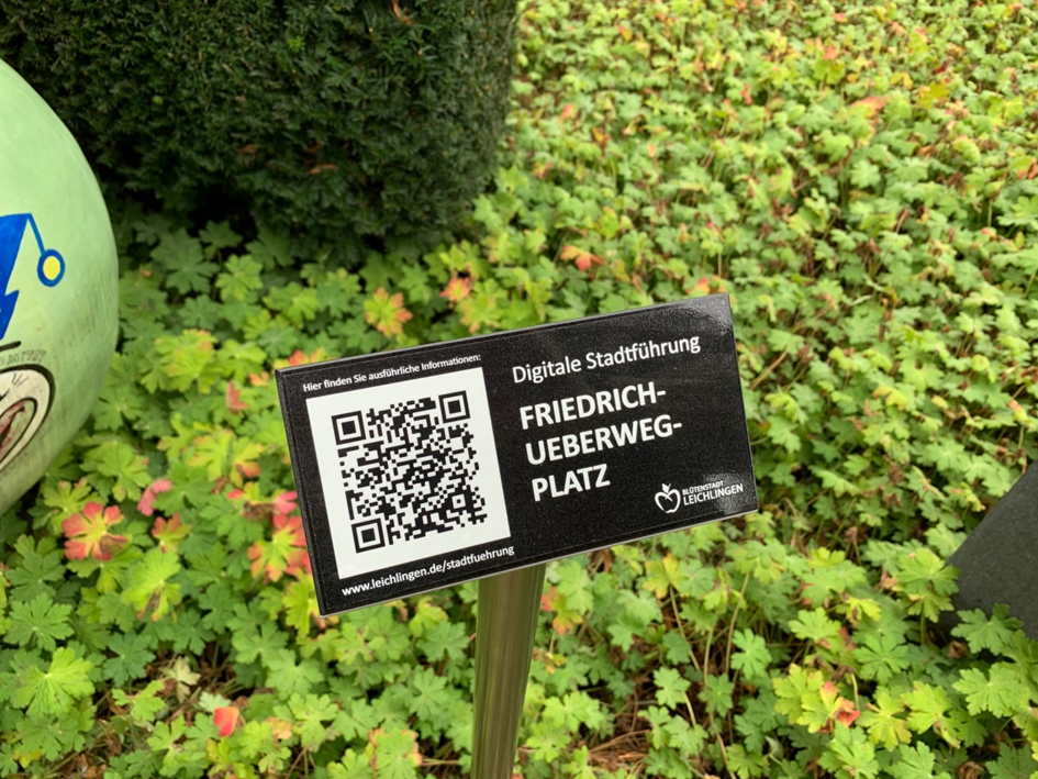 Eine Metallstele mit schwarzem Stein und der Aufschrift Friedrich-Überweg-Platz und QR-Code in weißer Schrift